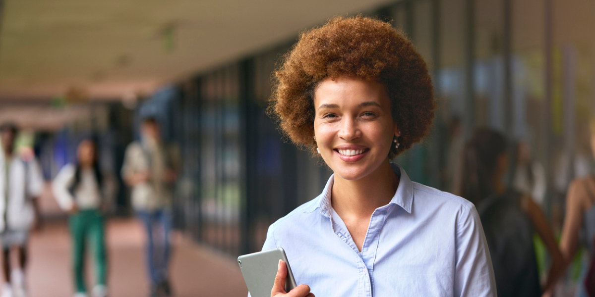 mulher negra com cabelo black power ruivo, camisa de botão azul segurando um tablet, em um ambiente aberto de escola.